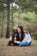 Naklejka na ściany i meble Young caucasian woman hugging her american staffordshire and french bulldog mixed breed dog at a pine forest.