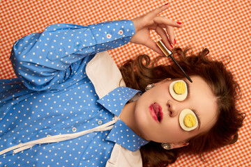 Creative makeup. Young girl lying on table with eggs on her eyes over peach color background. Vintage, retro style. Food pop art photography.