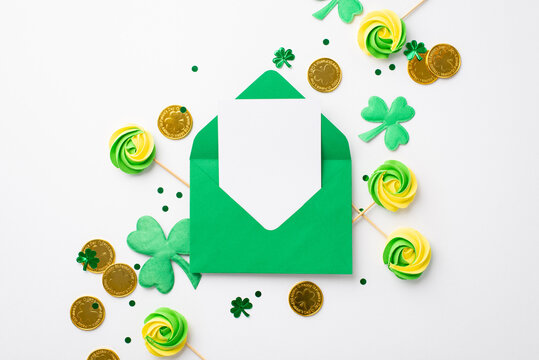 Saint Patrick's Day concept. Top view photo of open green envelope with paper card meringue candies gold coins trefoils and confetti on isolated white background with copyspace