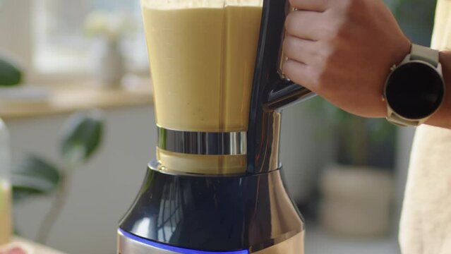 Tilt down close up shot of hands of unrecognizable man holding blender while preparing fruit smoothie in kitchen at home