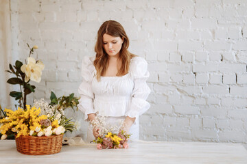 A young female florist in white clothes works against the background of a white brick wall, making bouquets in a flower shop. Workplace of a flower shop employee.