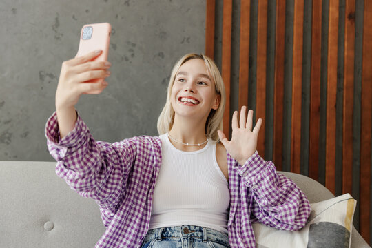 Young woman wear casual clothes do selfie shot on mobile cell phone waving hand sits on grey sofa couch stay at home hotel flat rest relax spend free time in living room indoor. People lounge concept.