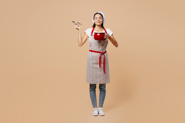 Full body young housewife housekeeper chef baker latin woman wear apron toque hat sniff smell scent of food close eyes hold pot open lid isolated on plain pastel beige background. Cook food concept.