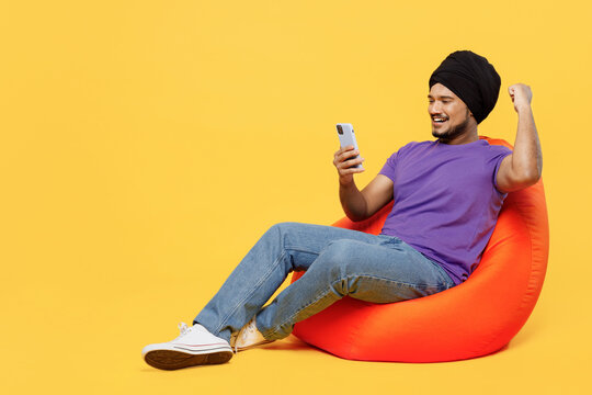 Full body devotee Sikh Indian man ties his traditional turban dastar wear purple t-shirt sit in bag chair use mobile cell phone do winner gesture isolated on plain yellow background studio portrait. - Powered by Adobe