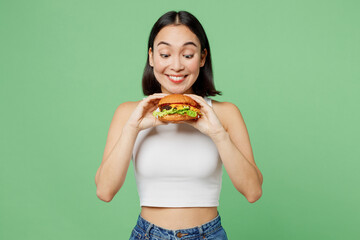 Young smiling happy excited woman wearing white clothes holding eating biting tasty burger isolated...