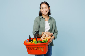 Plakat Young smiling woman wear casual clothes hold red basket with food products for preparing dinner look camera isolated on plain blue background studio portrait. Delivery service from shop or restaurant