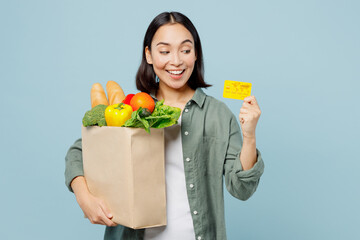 Fototapeta na wymiar Young satisfied fun woman wear casual clothes hold brown paper bag with food products credit bank card isolated on plain blue cyan background studio portrait. Delivery service from shop or restaurant