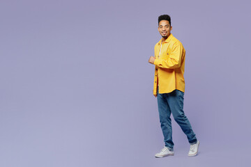 Full body sideways fun young man of African American ethnicity wear yellow shirt t-shirt walk go look camera isolated on plain pastel light purple background studio portrait. People lifestyle concept.