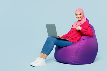 Full body young arabian muslim IT woman wear pink abaya hijab sit in bag chair work hold use point on show laptop pc computer isolated on plain blue cyan background People uae islam religious concept.