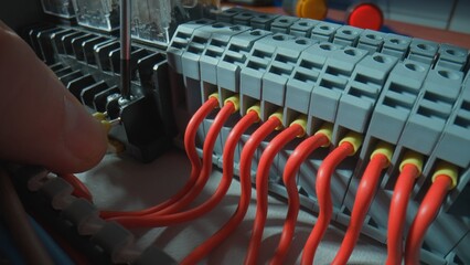 Gray plastic electrical panel with many red wires, yellow bushing ferrules, electrical parts, automatic switches. Close up of hand fasten the wire with a screw using a screwdriver.