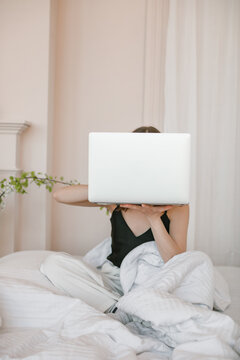 Woman sitting in bed holding a laptop computer in front of her face