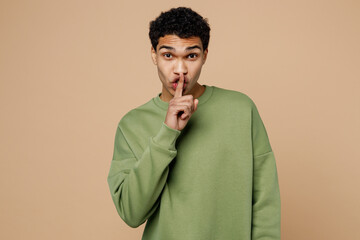 Young secret man of African American ethnicity wear green sweatshirt say hush be quiet with finger...