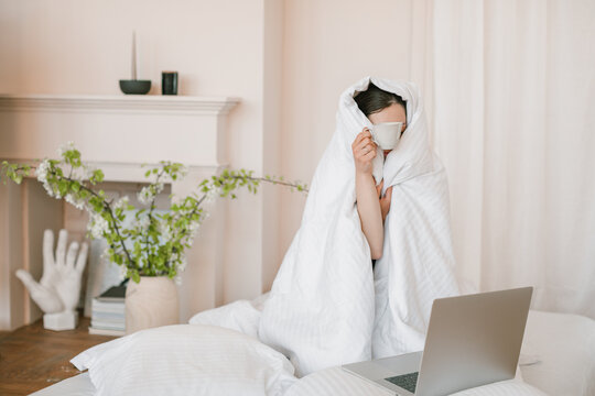 Woman wrapped in a duvet sitting in bed in front of a laptop computer drinking a cup of coffee