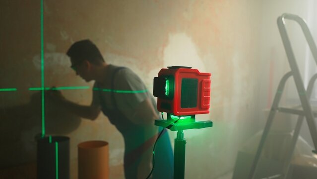 Green beam of laser roulette is displayed on brown wall. Male builder in blue construction overalls makes mark with pencil on wall at level of laser strip. Room under renovation. Soft light.