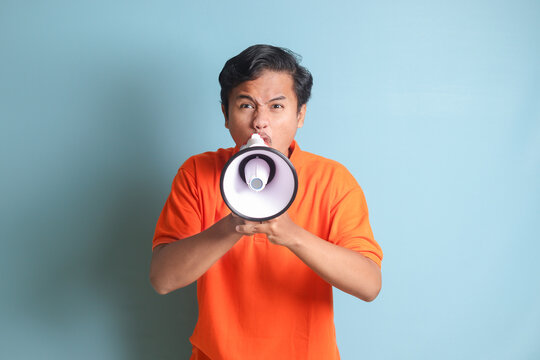 Portrait of attractive Asian man in orange shirt speaking louder using megaphone, promoting product. Advertising concept. Isolated image on blue background