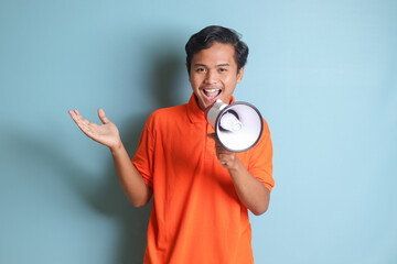 Portrait of attractive Asian man in orange shirt speaking louder using megaphone, promoting...