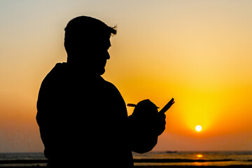 Silhouette of a man standing by the sea in twilight light during evening sunset. Silhouette view of a person writing something in a notebook.