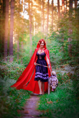 Red-haired beautiful girl in the forest. Girl from a fairy tale. The girl is walking with a wolf, a dog. Redhead. Outdoors. Girl in a red cape. Girl in a red coat. Summer. Little Red Riding Hood. Girl