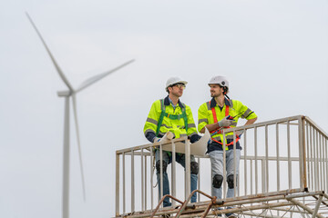 Naklejka na ściany i meble Two engineers in uniform and helmet discussing inspection and maintenance of wind turbine in wind farms to generate electrical energy, Renewable power energy