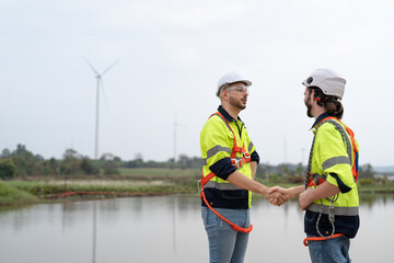 Two male engineers windmill in uniform and helmet safety shaking hands after to work success in wind turbine farm to generate electrical energy, Renewable power energy.