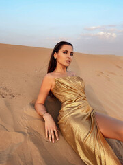 beautiful sexy woman with dark hair in elegant gold dress posing  in the desert at sunset