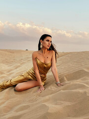 beautiful sexy woman with dark hair in elegant gold dress posing  in the desert at sunset