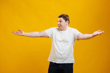 Handsome positive man wearing white t-shirt spread his hands to the sides and doesn't know what to do standing over yellow studio background