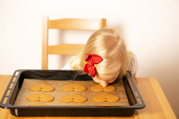 A little girl is sitting at the table with many cookies on the baked tray and smelling aromatic handmade heart cookies. Valentines Day or Mothers Day.