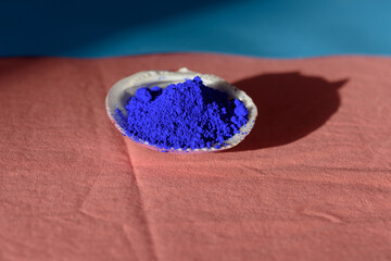Bodegón formado por una concha llena de pigmento azul cobalto sobre tela rojiza y franja superior azulada, y proyección de sombra lateral. - obrazy, fototapety, plakaty