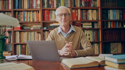 Portrait of friendly senior man working on laptop in library, doing research