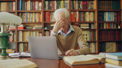 Senior man translator rubbing tired eyes, working long hours on laptop, writer