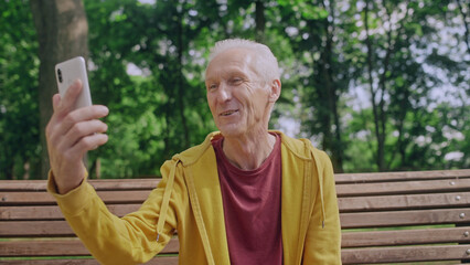 Mature man making video call to friends or relatives, sitting on bench in park
