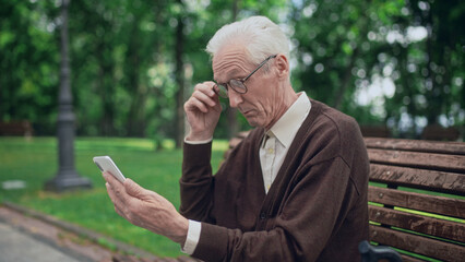 Elderly man reading news on smartphone through eyeglasses sitting on bench, bad eyesight