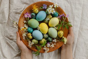 Obraz na płótnie Canvas Stylish easter eggs and blooming spring flowers in wooden bowl in woman hands. Happy Easter! Rustic easter flat lay. Natural painted eggs and blossoms on rural table