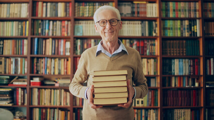 Senior male writer holding a stack of books at the library and smiling, doing research
