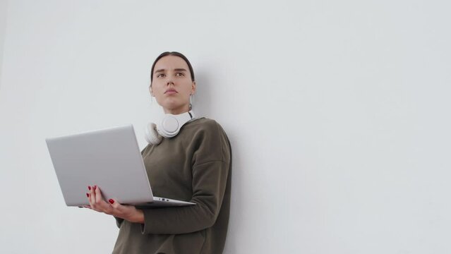 Modern young female IT specialist standing against white wall background working on laptop