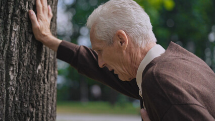 Anxious senor man feeling pain in chest, leaning on tree and breathing heavily, needs help