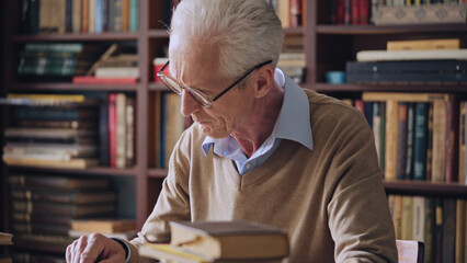 Senior male in eyeglasses reading books in the library, scientist doing research