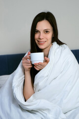 Attractive brunette woman wrapped in a white blanket smiling with cup of tea or coffee in the morning, vertical video. She is sitting on bed and looking at camera. Lazy morning, good mood concept.