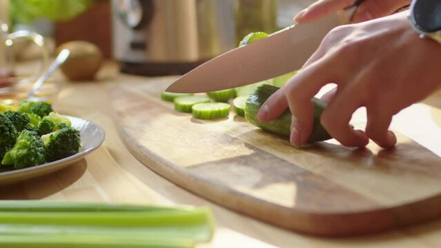 Close up shot of hands of man cutting fresh cucumber on wooden board at kitchen table while preparing healthy meal at home