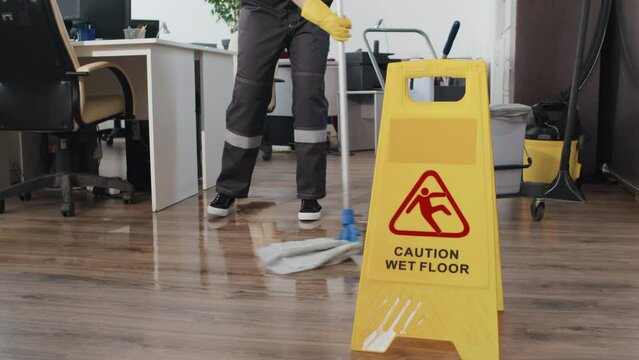 Selective focus slowmo of yellow plastic signboard with caution warning about slippery wet floor, unrecognizable cleaner using mop on background