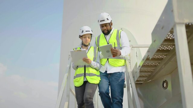 Two engineers african american man and woman in uniform walking down the stairs talking and use tablet to verify that a wind turbine is running correctly and effectively, Renewable energy