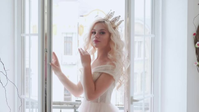Portrait fantasy blonde glamour woman princess stands near panoramic huge window white classic room interior. Fashion model girl posing looking at camera pretty beautiful face. Pink royal luxury dress