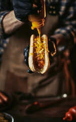 A guy in a leather apron is preparing a hot dog. The chef in gloves pours mustard over a hot dog. Creation of fast food.