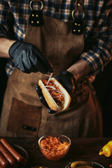 A guy in a leather apron makes a hot dog. A chef in black gloves puts an onion in a bun.
