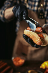 A guy in a leather apron makes a hot dog. The chef in black gloves puts a sausage in a bun.