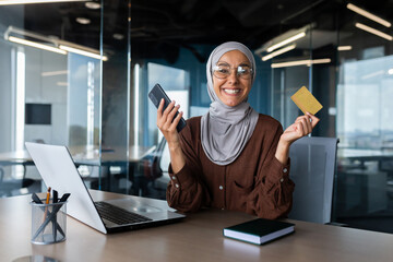 A young Arab woman in a hijab works in the office at a desk. He is holding a credit card and a phone. Happy smiling at the camera.