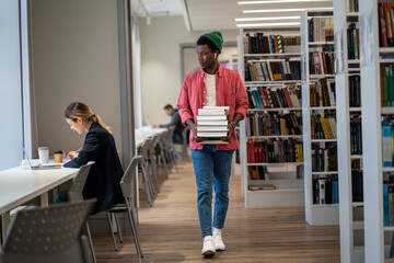 Fototapeta na wymiar Bookworm. Full length of African American international student guy walking with stacked books through college library. Male librarian organizing and managing resources. Education concept