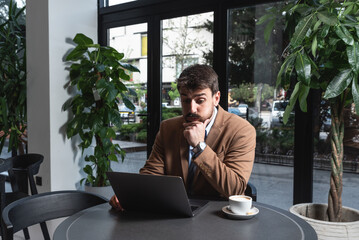 Young stressed nervous annoyed businessman sitting in cafeteria restaurant drinking coffee reluctantly looking at laptop computer monitor, unhappy and upset because of bad business decision