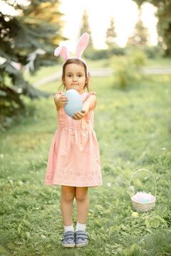 Little cute girl wear bunny ears holding big blue painted egg on Easter egg hunt in park